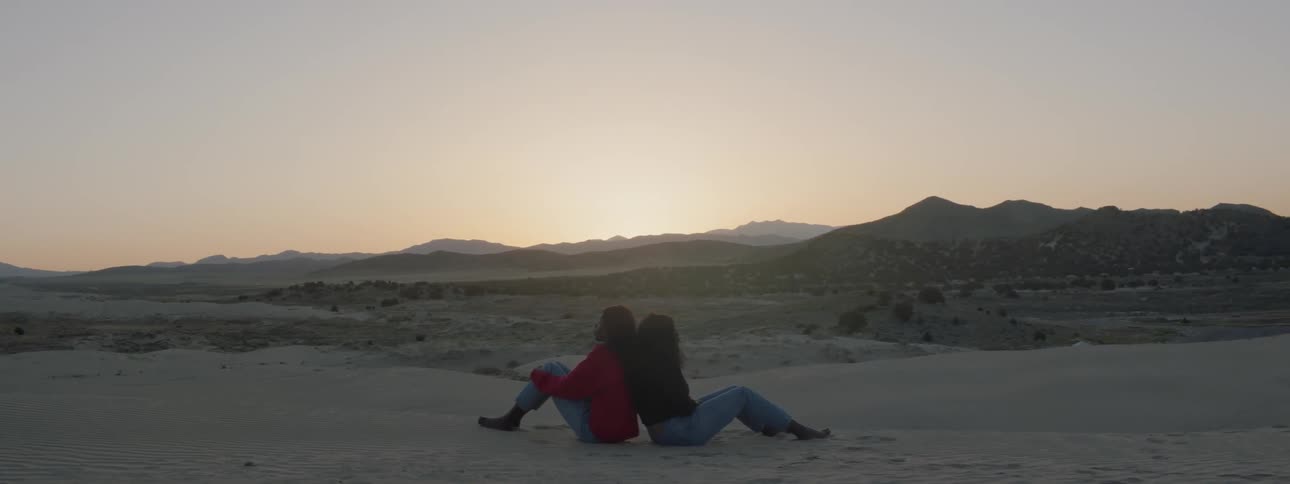 Two women sitting back to back in the desert at dusk