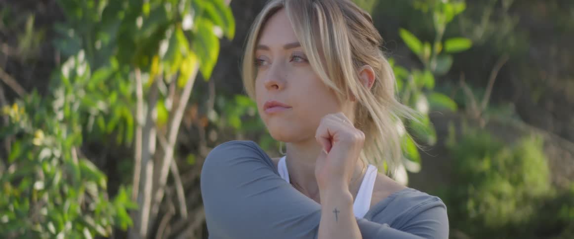 A blonde haired woman crosses her arms to stretch as she stands in front of a forest