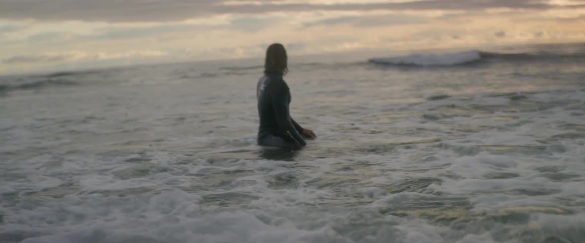 A man sits on his surfboard in the ocean and looks over the water at the sunset