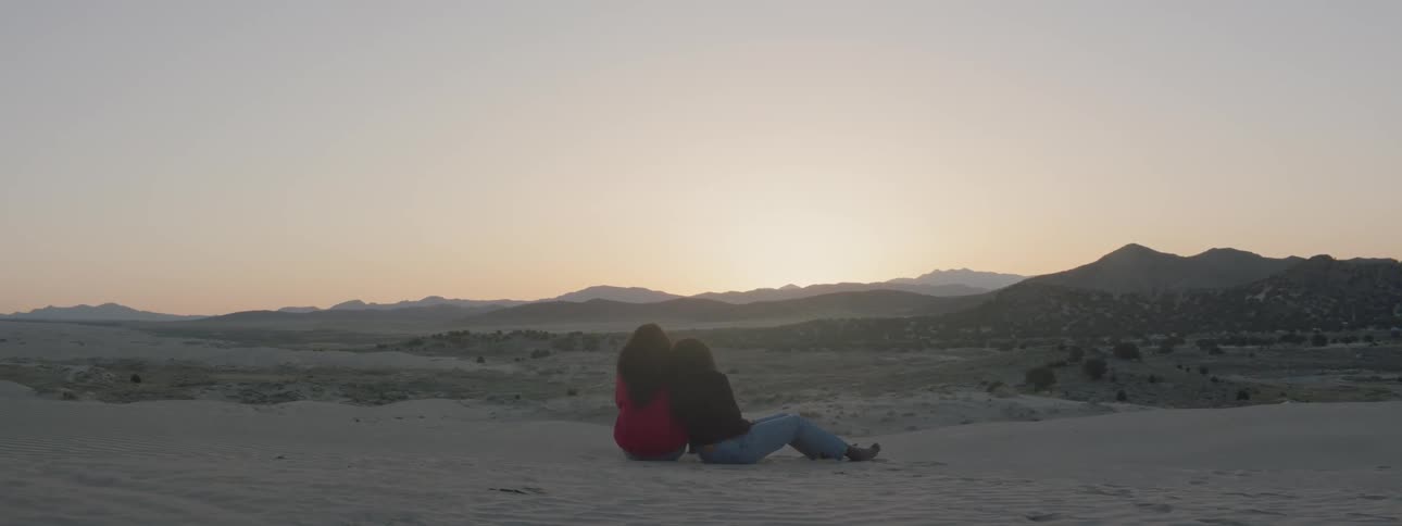 Two women sit in the desert at sunset