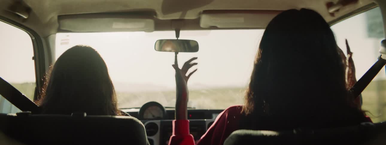 Two girls sing and dance while driving through a remote area in a car