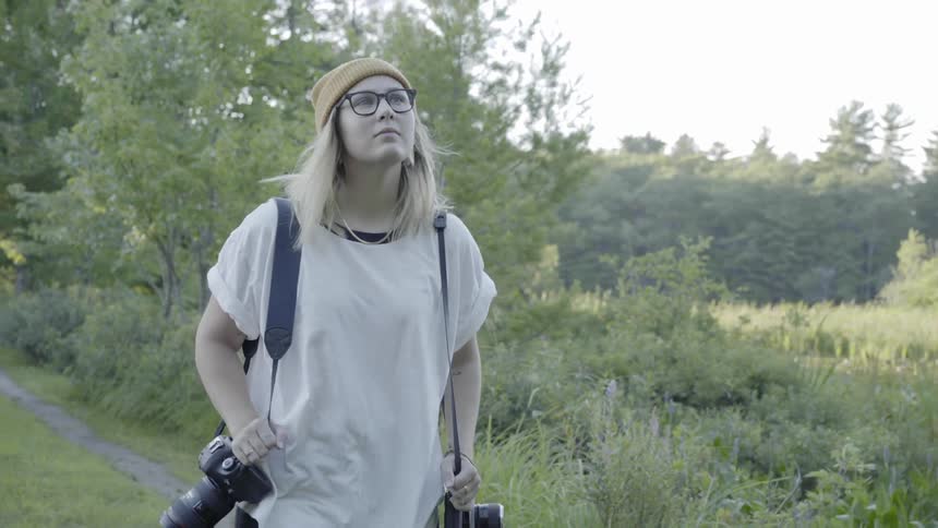 A woman walks down a path created by vehicles in the woods with cameras on her shoulders