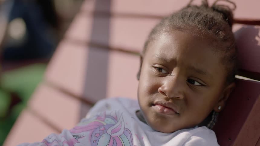 A little girl sits in a chair frowning while another child plays near her