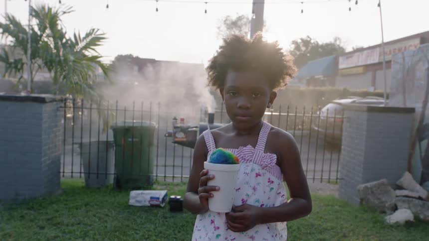 A young girl holds a snow cone in a glass next to a grill