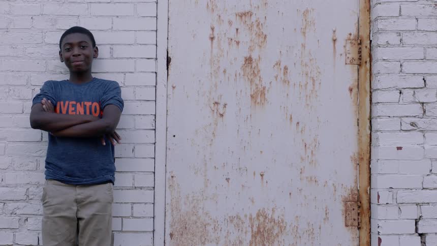 A boy stands smiling with his back to a wall as he crosses his arms