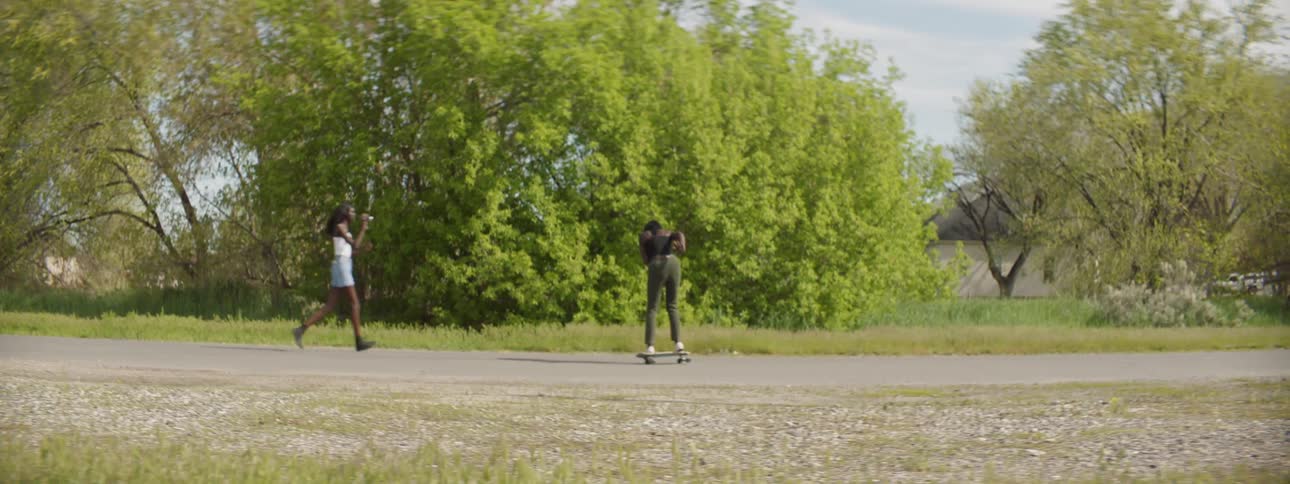 A woman rides by on a skateboard while another woman runs after her filing her