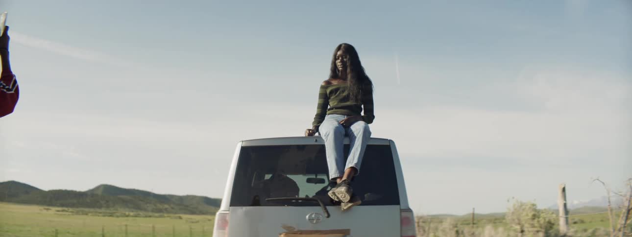 A woman sitting on a car adjusts her hair so another woman can photograph her