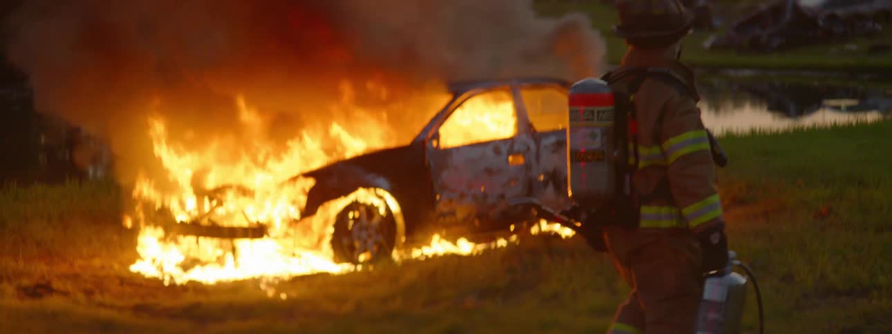 A firefighter walking up to a car that is burning in a field