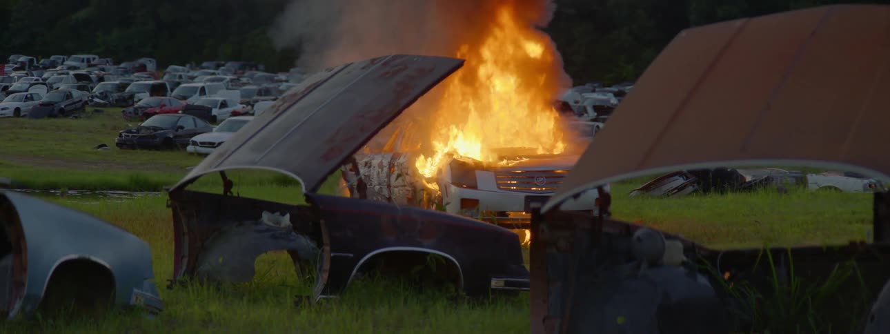 A fire blazes on a car in a junk yard full of cars