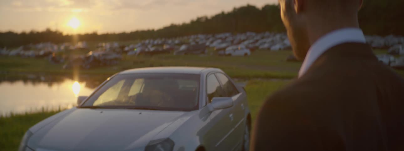 A man in a suit that is looking at some cars that are in a field