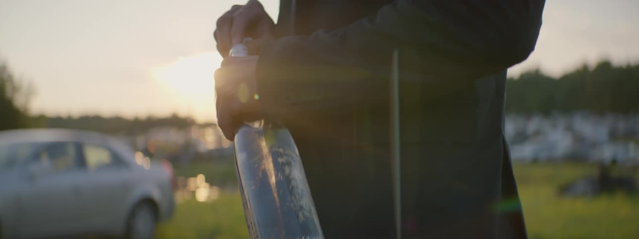 A man standing in a grassy parking lot at sunset holding a bottle and putting a rag in it