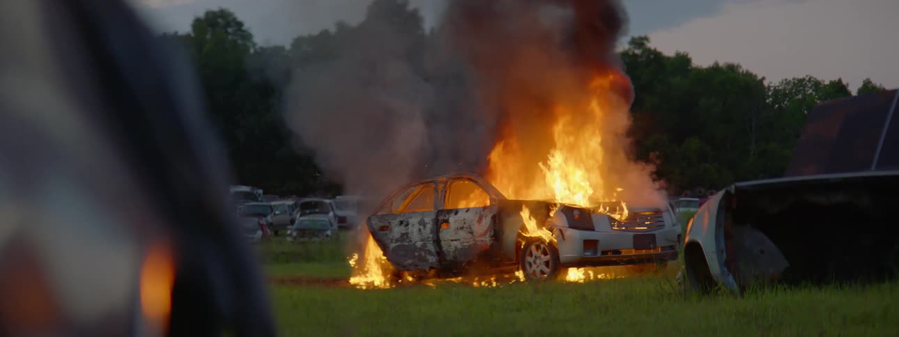 A car that is on fire in the middle of a scrap yard field