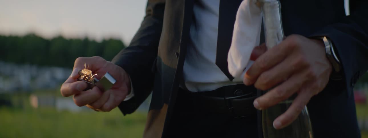 A man pulls a lighter out of his tuxedo jacket and moves the lighter toward a molotov cocktail he is holding in his other hand