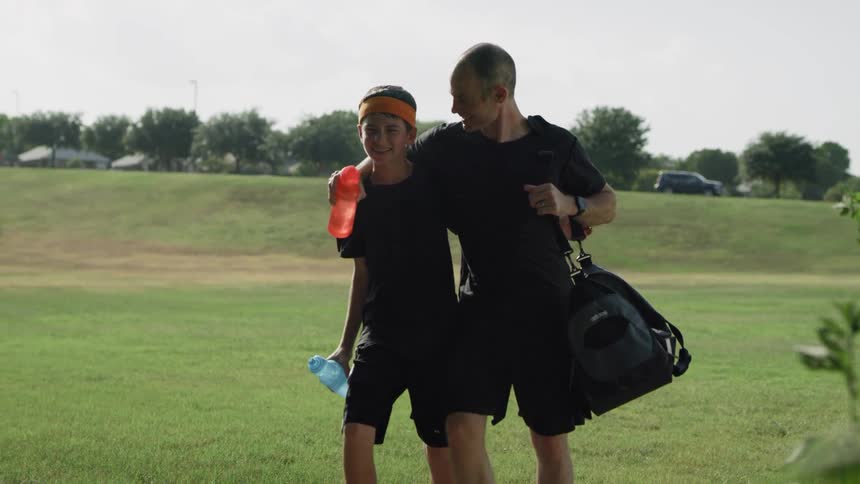 A man carries a sports bag and a water bottle and puts his arm around a boy holding a water bottle