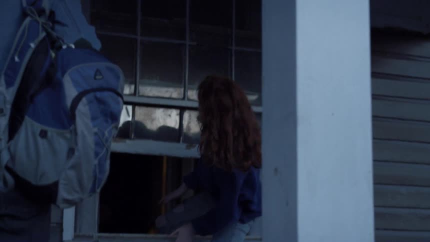 A young woman climbs through a window as a young man holding a backpack waits by the door