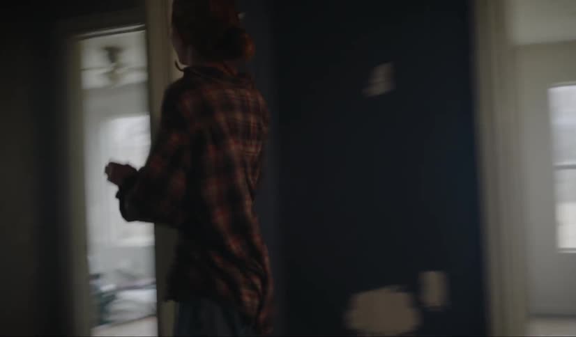 A woman looks out the window of a dark room then runs to another room in the house