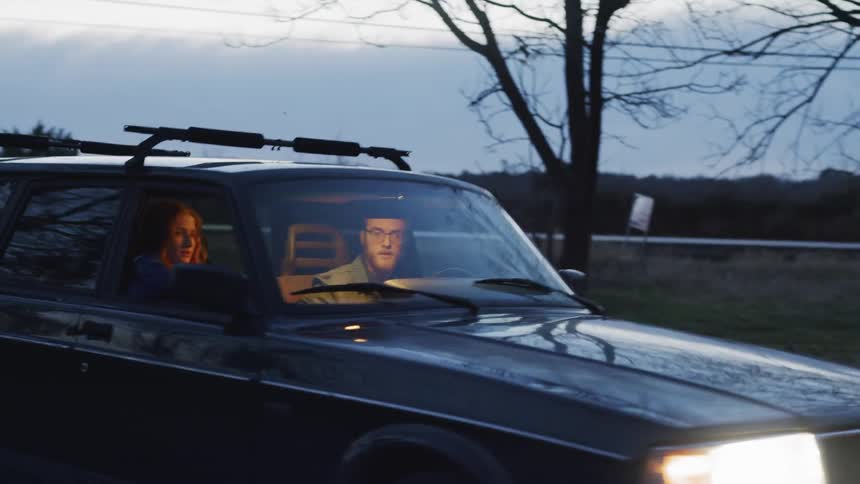 A man drives a car at dusk with a woman in the passenger seat and parks it