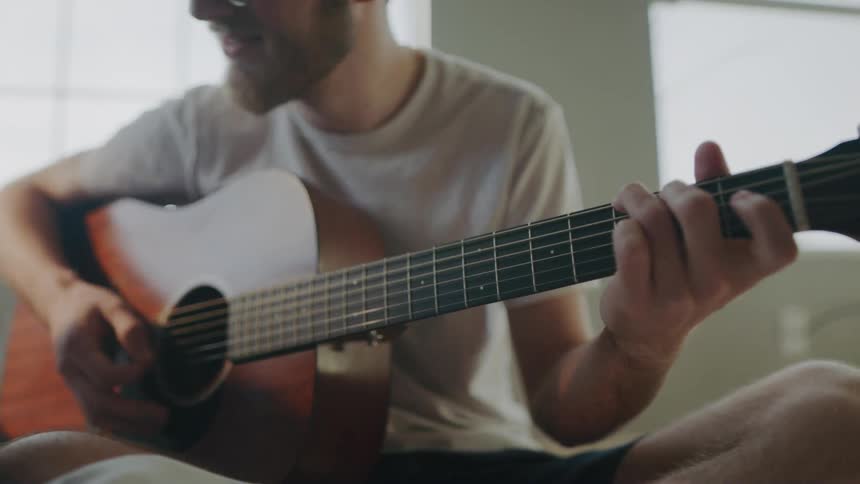 A man sitting on a bed strumming a guitar
