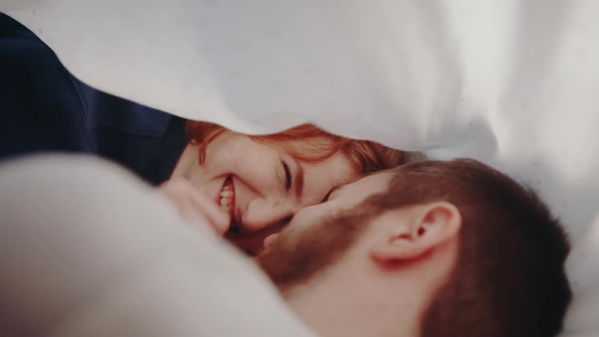 A man and a woman look at each other and laugh under a bed sheet
