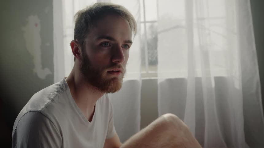 A man in a white shirt looks perplexed as he sits on the floor in front of a window