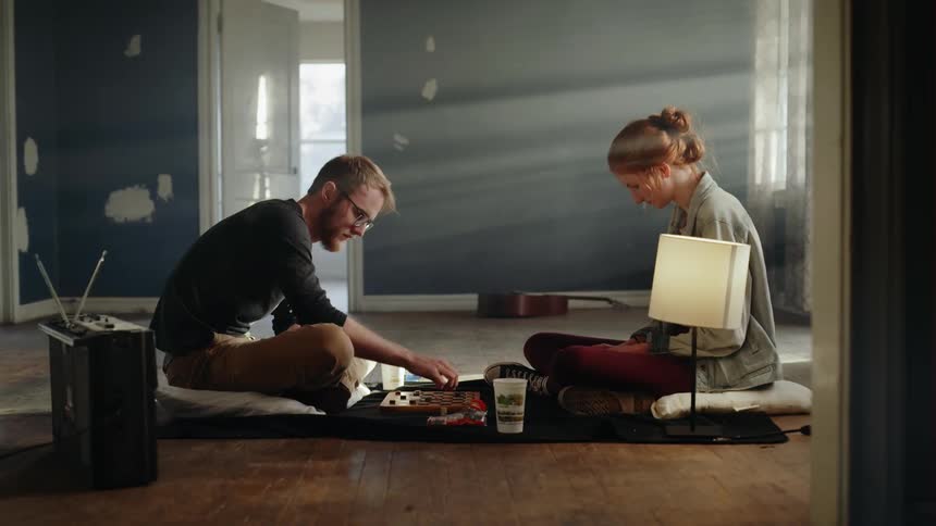 A man and a woman play checkers on a blanket on the floor of an unfurnished room