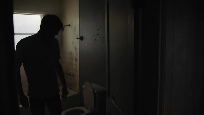 A man stands in a dark dank bathroom then walks into a cleaner brighter room