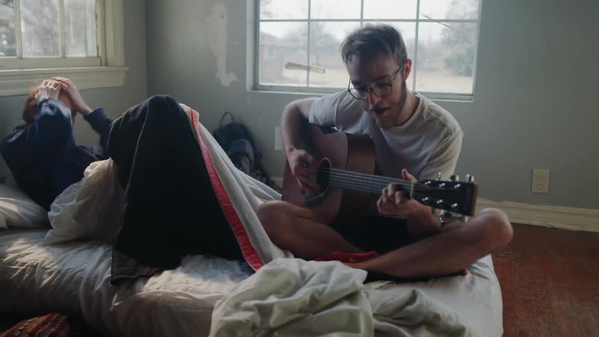 A man playing the guitar in bed for a woman laughing hysterically