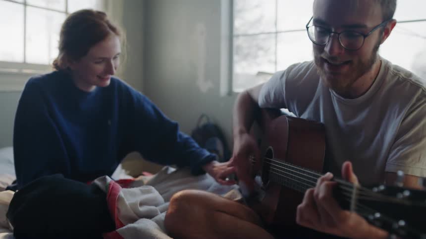 A man playing a guitar on a bed for a woman