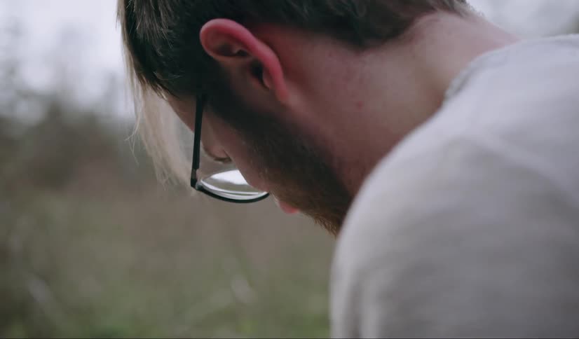 A caucasian man looks down at something he is holding and sitting down with glasses on his face and short facial hair