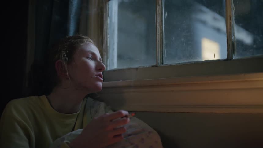 A person sitting close to a window smoking a cigarette and blowing the smoke out