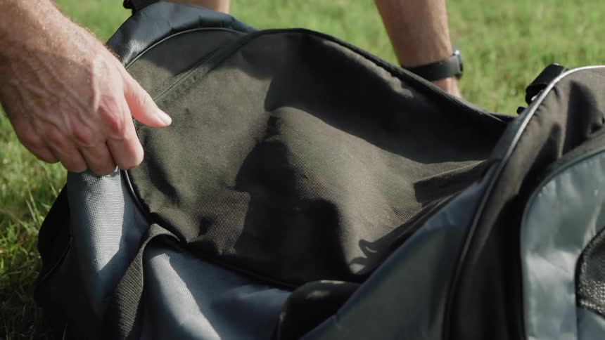A man zips closed a bag laying on the grass then picks it up