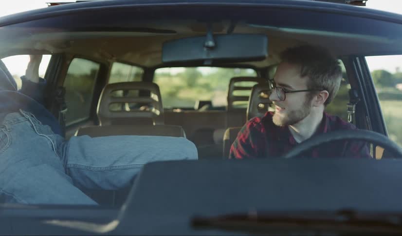 A woman with red hair sits in the passenger seat next to a man smoking a cigarette until she leans out the window then gets back in