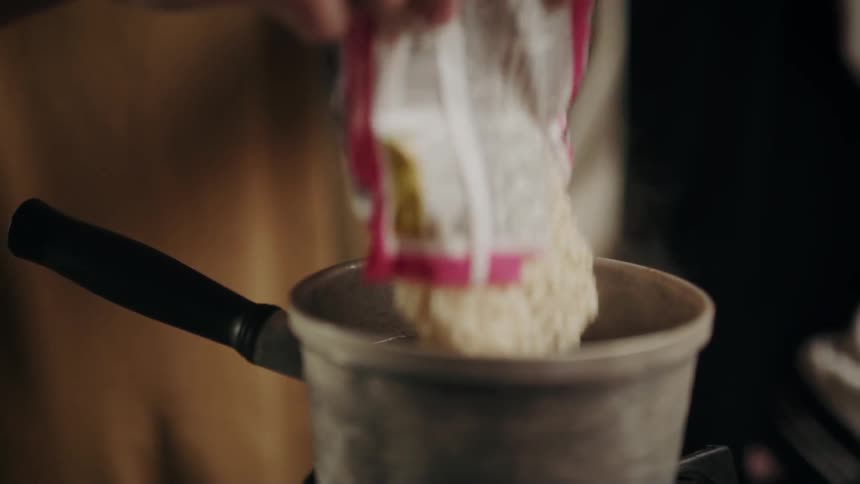 A person dumps top ramen into hot water in a pan on the stove