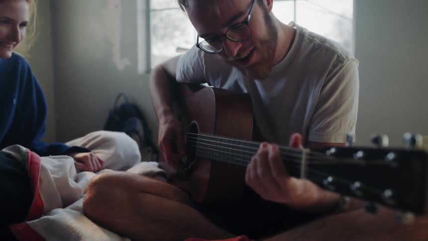A woman laughs while a man plays a guitar in the bed they are sitting on