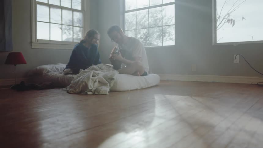 A man and woman sitting on a mattress while the man plays guitar