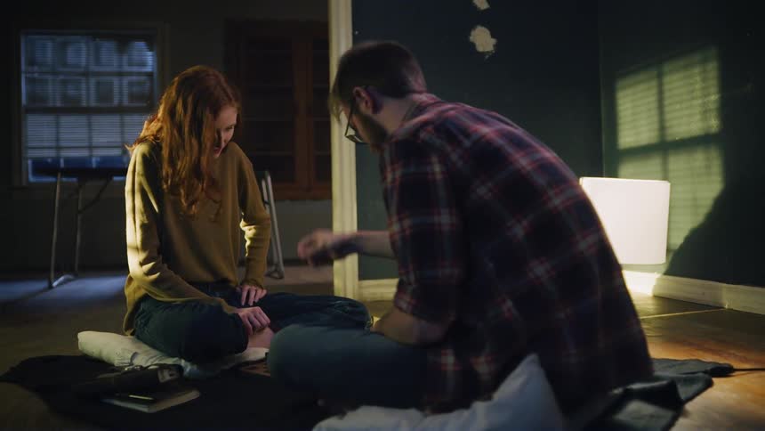 A man and a woman sit on cushions on the floor and talk