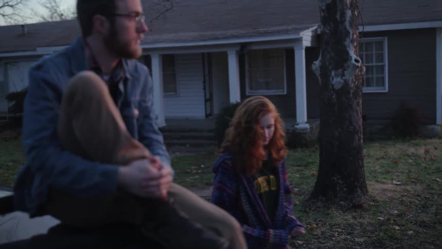 Man sitting on a car in the front yard of a house while a woman walks up to him
