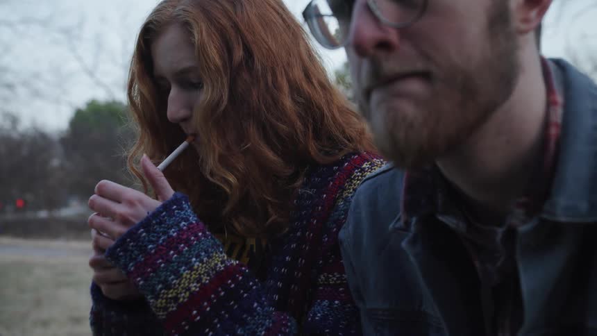 Woman smokes cigarette while sitting next to man outdoors