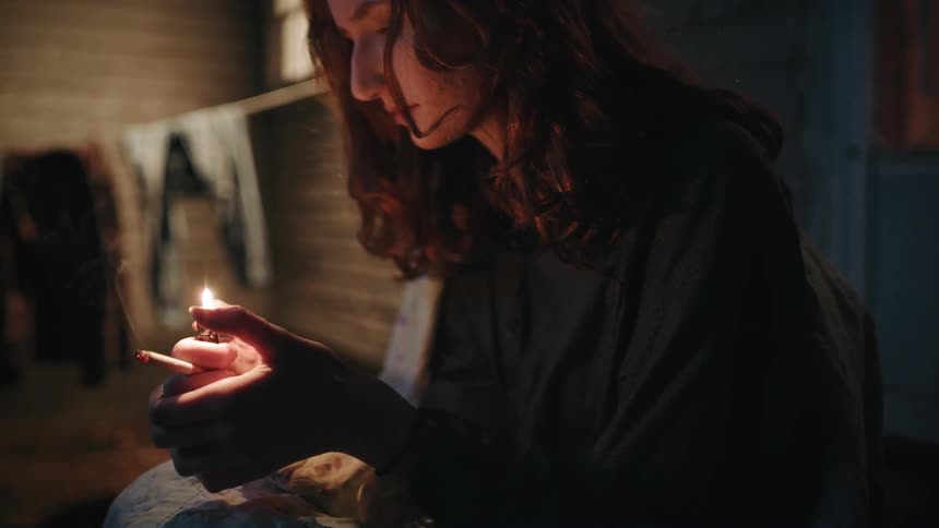 A woman sits outside at night holding a cigarette and a lighter