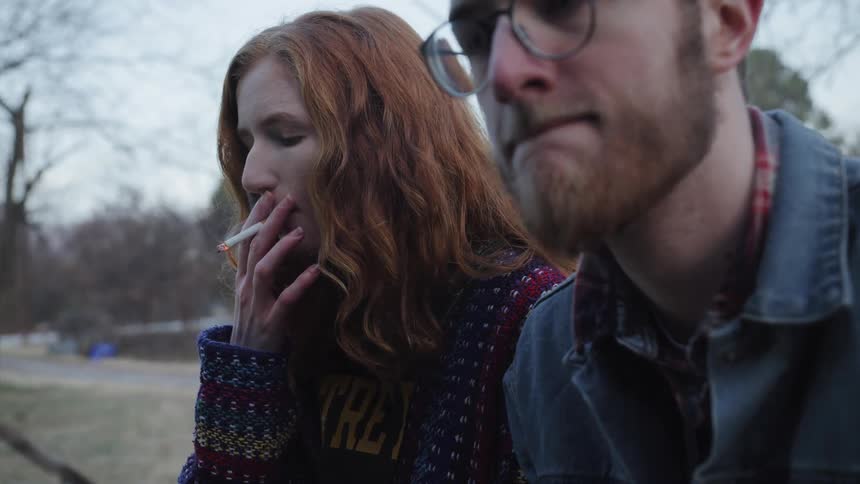 A woman smokes a cigarette while sitting outside next to a man