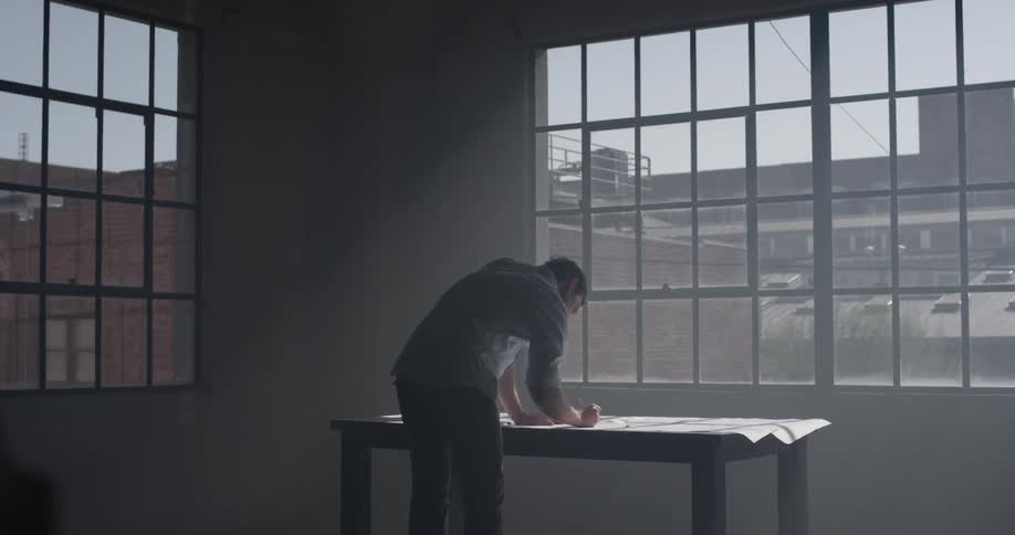 A man stands in front of a city window in the sunlight drawing on paper with a pencil