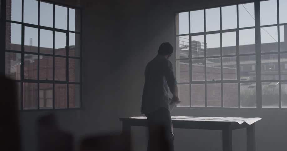A man that is standing at a desk in front of a window rolling up a piece of paper