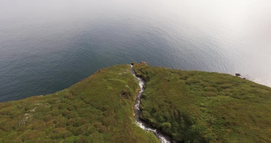 A stream winding along the top of a cliff and falling over the edge onto rocks and then into a large body of water