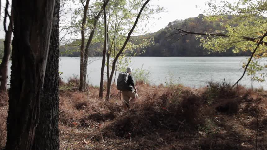 A young man wearing a large backpack and using walking sticks as he walks through brush towards a body of water