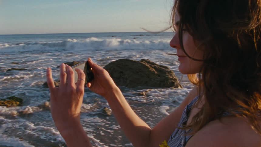 A woman stands on the beach trying to take a photo of the surf