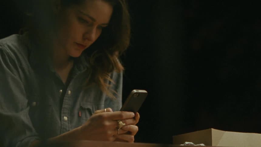 A young, brunette woman sitting while she types on her phone as a man eating out of a white bag stands nearby