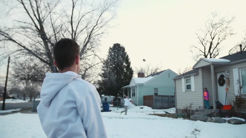 Two boys stand on their front lawn tossing the football is a snow while girl standing on a porch looks on