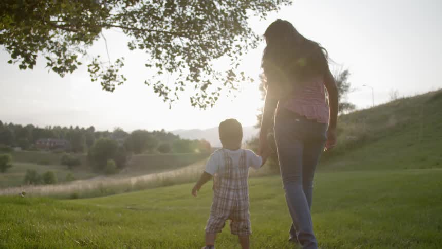 A mother holds her toddler's hand as they walk across a green hilltop at sunset