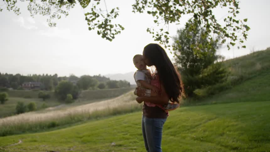 A woman holds a toddler and kisses him as they stand atop a green hill in late afternoon