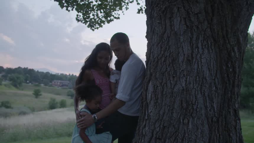 A man leans against a large tree and embraces his wife and two children